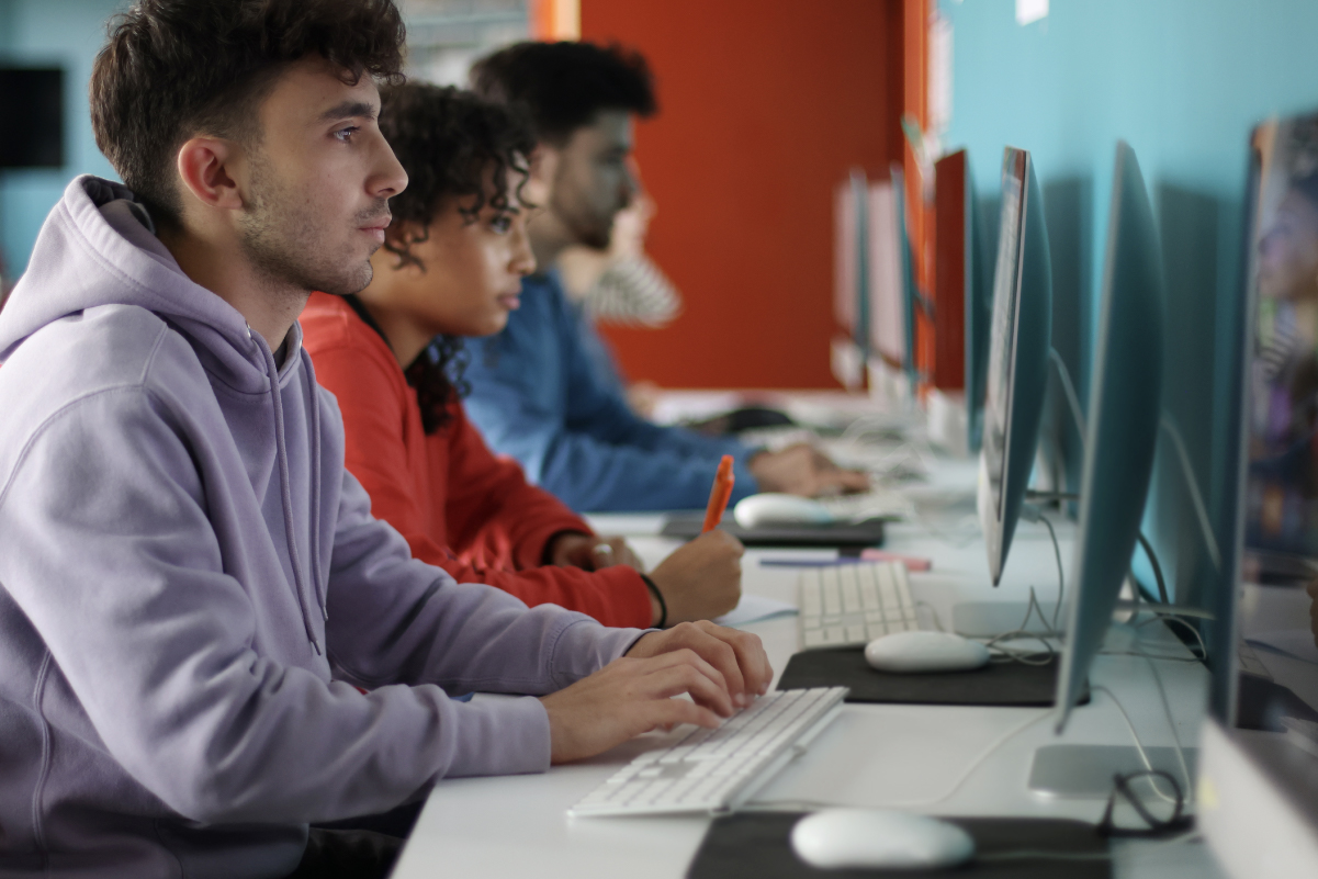 Giovani seduti in fila davanti a computer desktop in un'aula informatica; in primo piano un ragazzo con felpa viola digita mentre altri studenti concentrati lavorano ai monitor