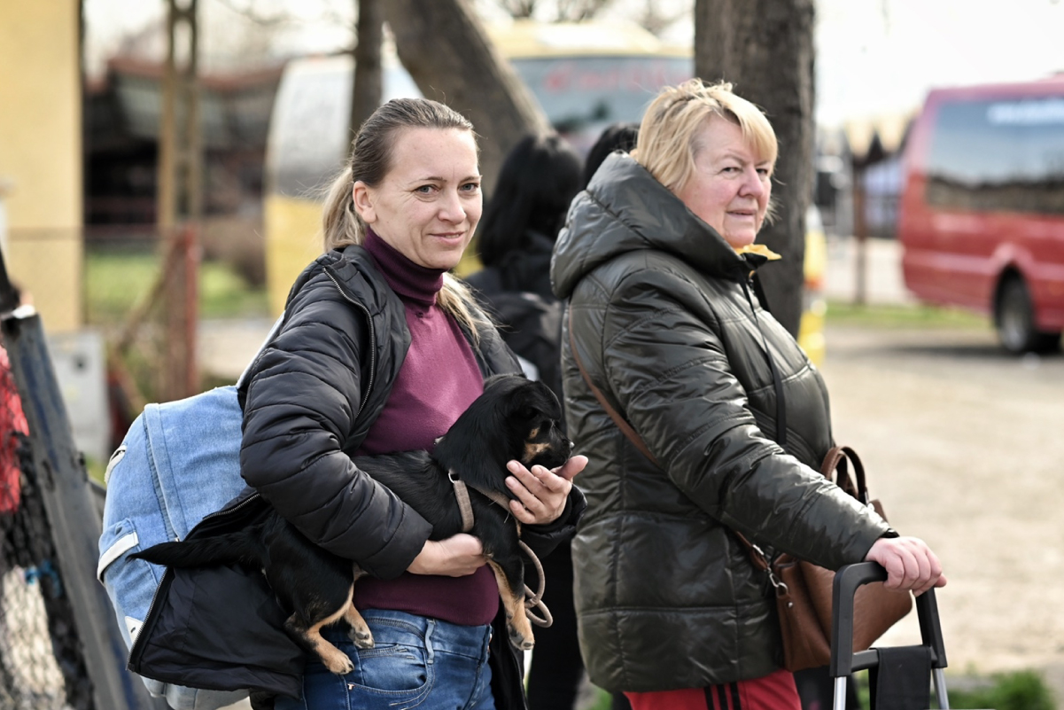 Due donne con giacche scure camminano all'aperto; una tiene in braccio un cane nero mentre l'altra trascina una valigia, con un autobus rosso sfocato sullo sfondo