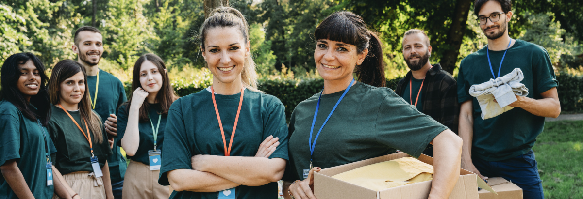 Gruppo di giovani volontari con magliette verdi e badge identificativi sorride all'aperto; una volontaria tiene un pacco mentre un volontario solleva una scatola, con alberi sullo sfondo