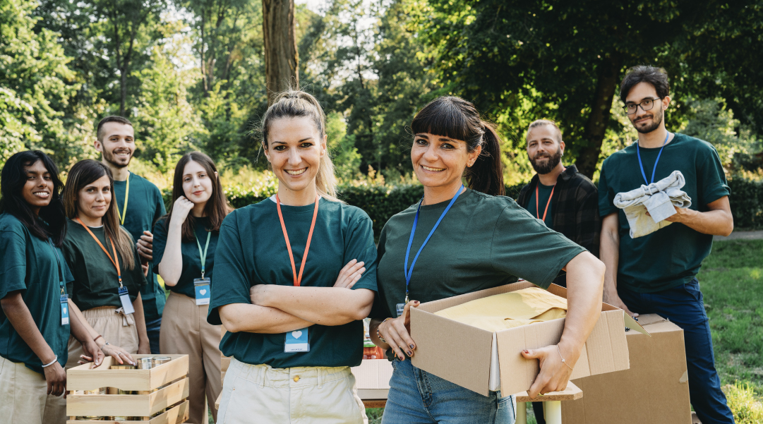 Volontarie e volontari con magliette verdi e badge si riuniscono all'aperto con scatole e cassette di cibo; una volontaria al centro incrocia le braccia mentre un ragazzo con occhiali regge un pacco