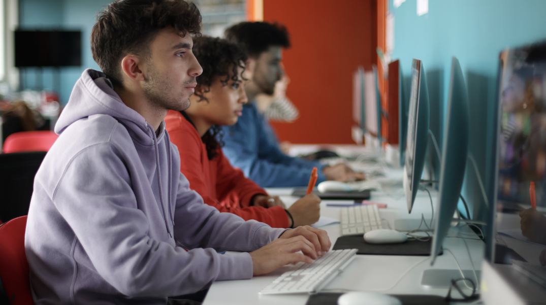 Ragazzo con felpa viola utilizza un computer in un'aula informatica con altri studenti seduti in fila davanti ai monitor