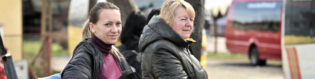 Dettaglio orizzontale di due donne con giacche scure che camminano all'aperto; una guarda la fotocamera mentre l'altra sorride, con un autobus rosso sfocato sullo sfondo
