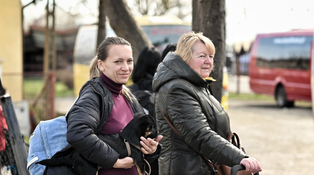 Due donne con giacche scure camminano all'aperto; una tiene in braccio un cane nero mentre l'altra sorride, con un autobus rosso e un campo sullo sfondo