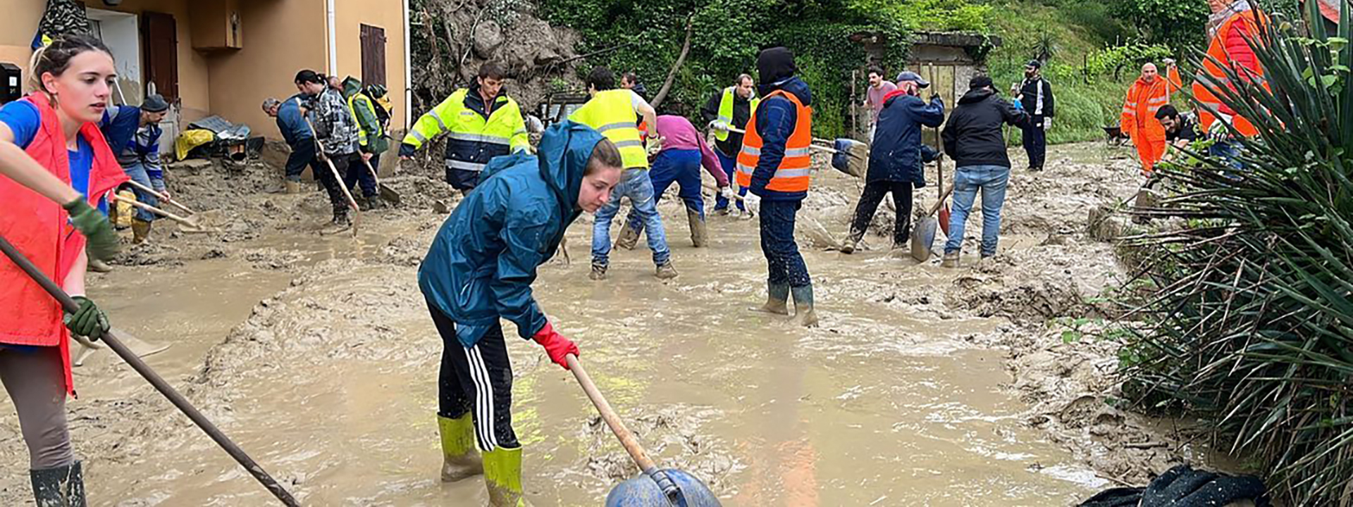 Volontari con stivali e pale spalano il fango in un cortile allagato dopo una frana, lavorando in gruppo con giubbotti ad alta visibilità