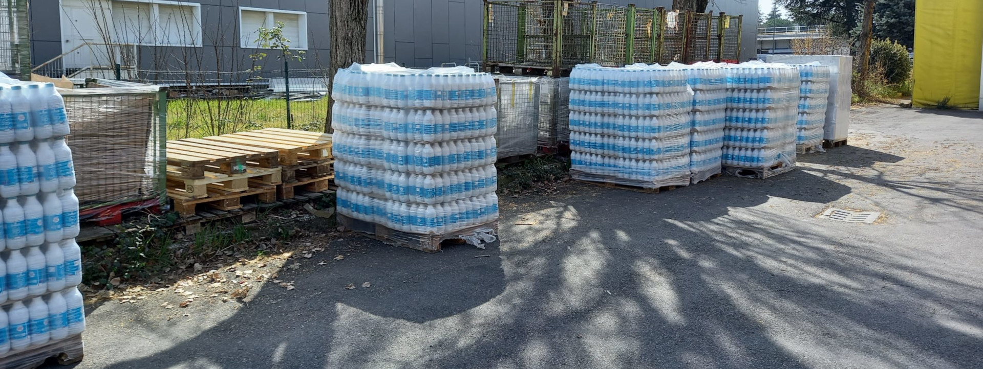 Pallet di bottiglie di latte con etichetta azzurra avvolti in pellicola trasparente all'esterno di un magazzino con bancali e cassoni sullo sfondo