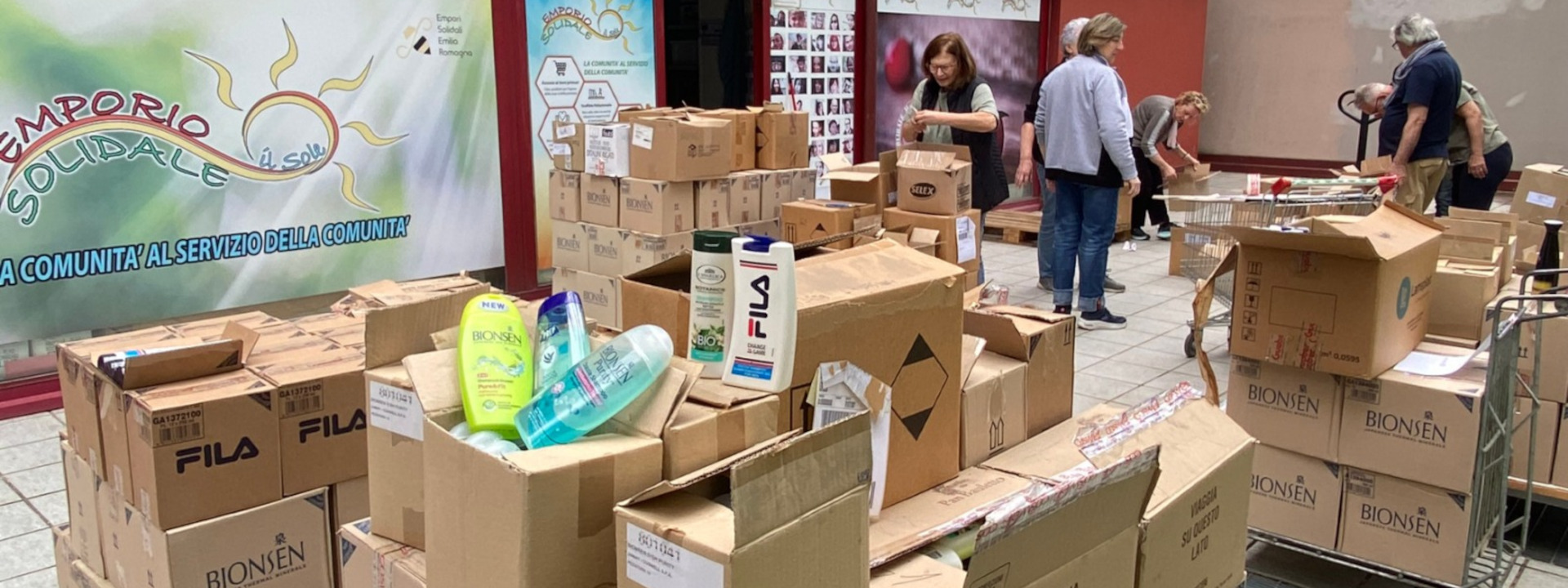 Area di raccolta donazioni con numerosi scatoloni di cartone e prodotti per la pulizia esposti in primo piano, e volontari che organizzano i pacchi davanti a un colorato striscione