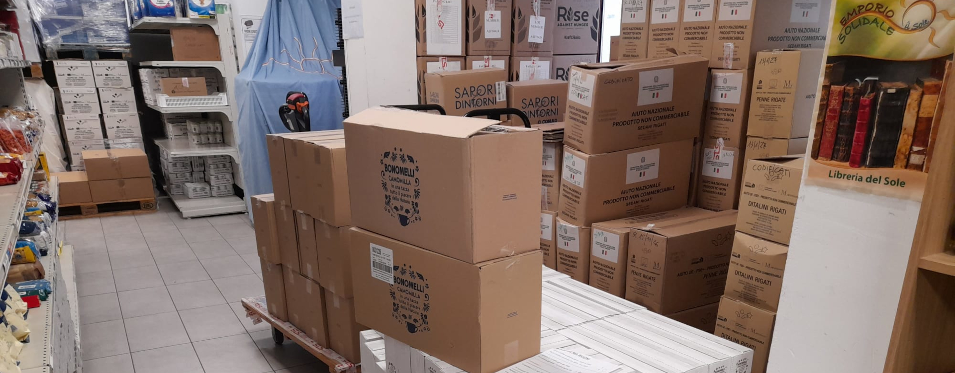 Pile di scatoloni imballati in un magazzino con scaffali e prodotti alimentari sullo sfondo, preparati per una donazione in Montenegro