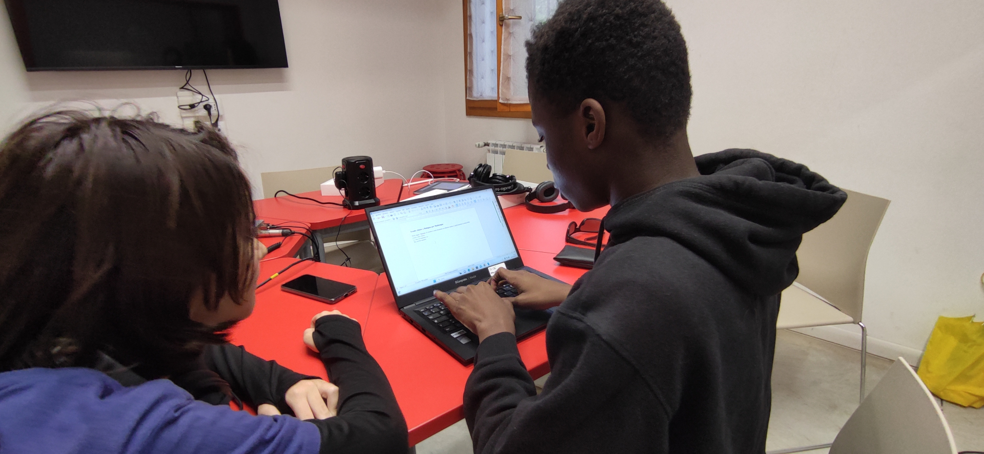 Due persone sedute a un tavolo rosso guardano lo schermo di un laptop durante un'attività formativa di donazione di computer