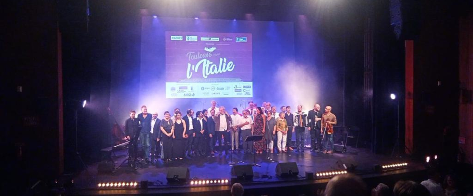 Fine concerto dell'evento "Toulouse pour l'Italie" con un grande gruppo di artisti e organizzatori sul palco illuminato e schermo con logo dell'evento sullo sfondo