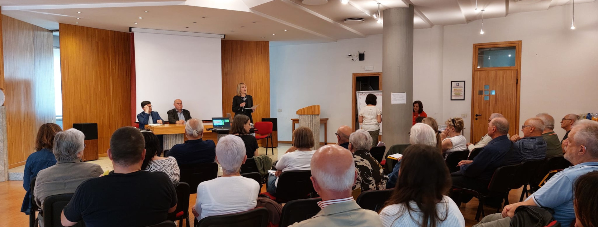 Partecipanti seduti assistono a un incontro formativo in una sala conferenze con tre relatori seduti a un tavolo e una relatrice in piedi che parla al pubblico