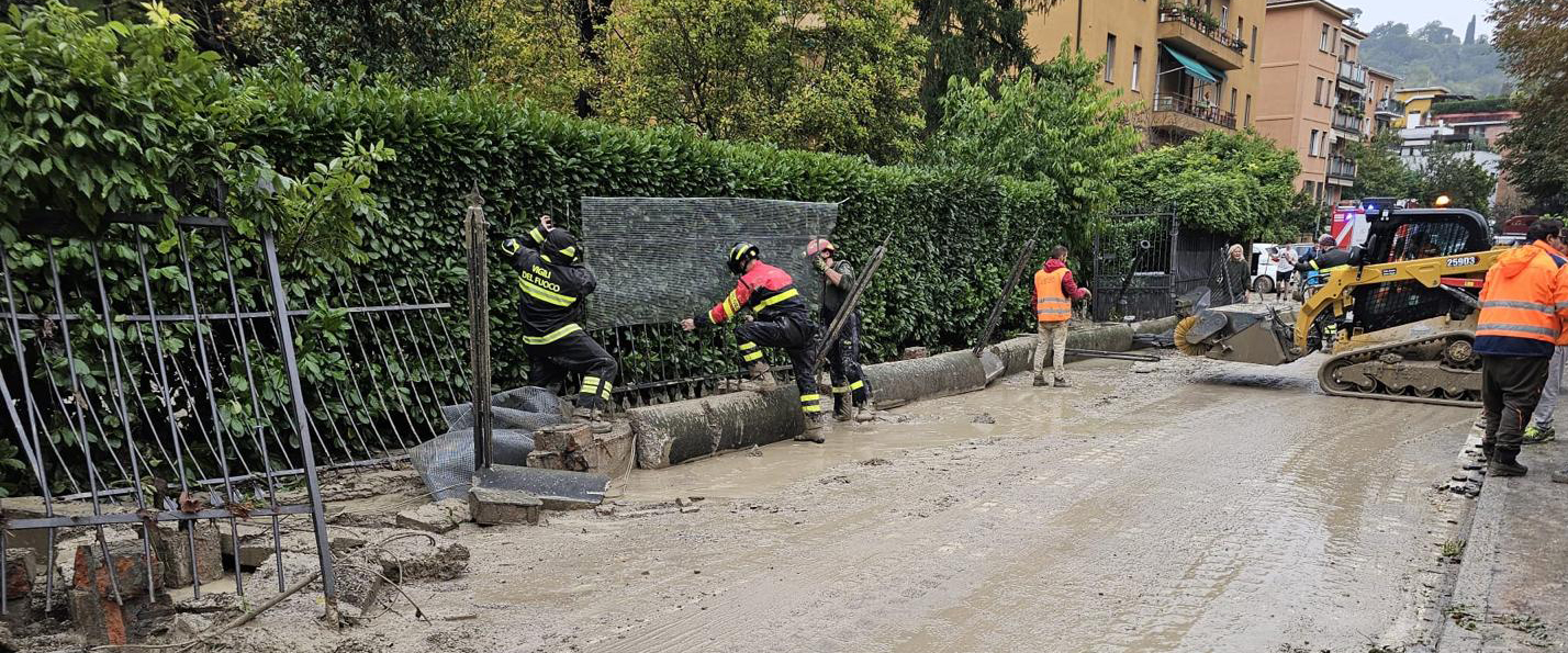 Operazioni di pulizia dopo un'alluvione: vigili del fuoco e operai rimuovono fango e macerie da una strada, con escavatore in azione e case sullo sfondo.