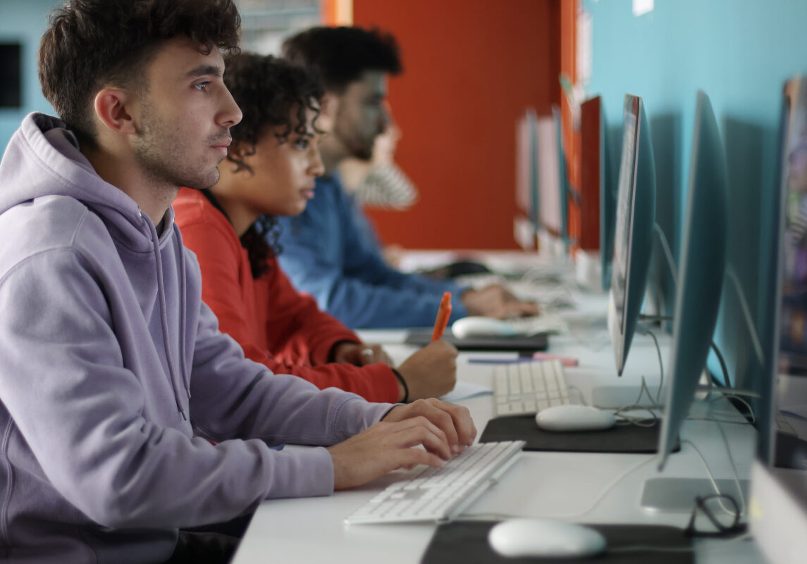Giovani seduti in fila davanti a computer desktop in un'aula informatica; in primo piano un ragazzo con felpa viola digita mentre altri studenti concentrati lavorano ai monitor