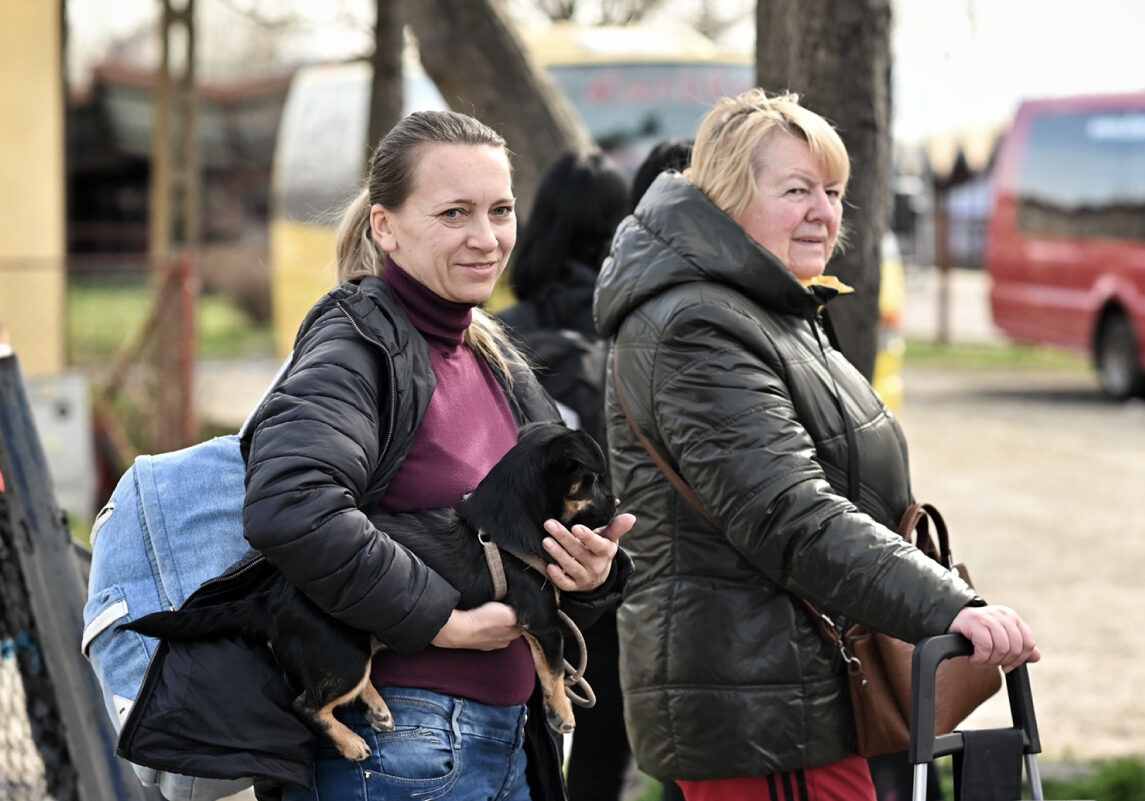 Due donne con giacche scure camminano all'aperto; una tiene in braccio un cane nero mentre l'altra trascina una valigia, con un autobus rosso sfocato sullo sfondo